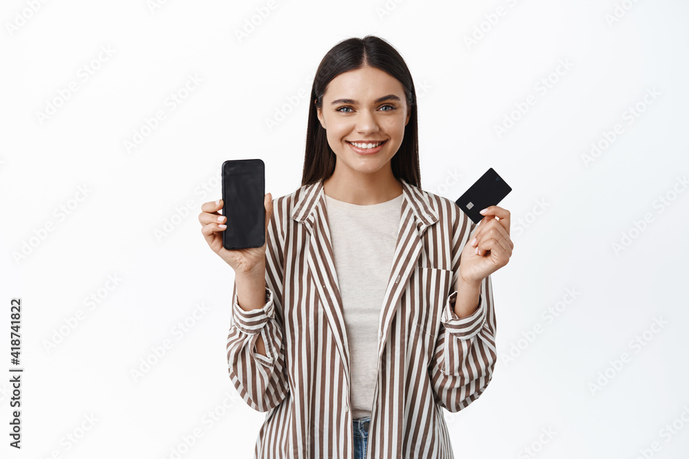 Portrait of modern young woman in stylish outfit, showing smartphone empty screen and plastic credit card, smiling pleased, recommending an app, white background Portrait of modern young woman in stylish outfit, showing smartphone empty screen and plastic credit card, smiling pleased, recommending an app, white background