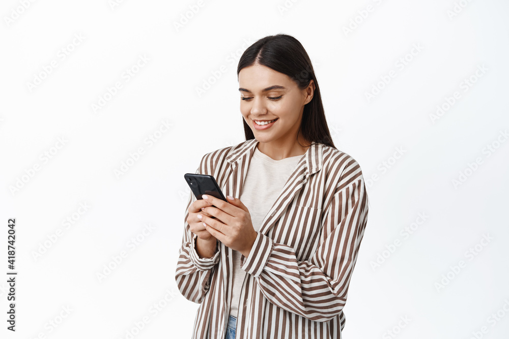 Young modern woman in stylish outfit using her mobile phone, chatting on smartphone app and smiling, reading screen, standing over white background Young modern woman in stylish outfit using her mobile phone, chatting on smartphone app and smiling, reading screen, standing over white background