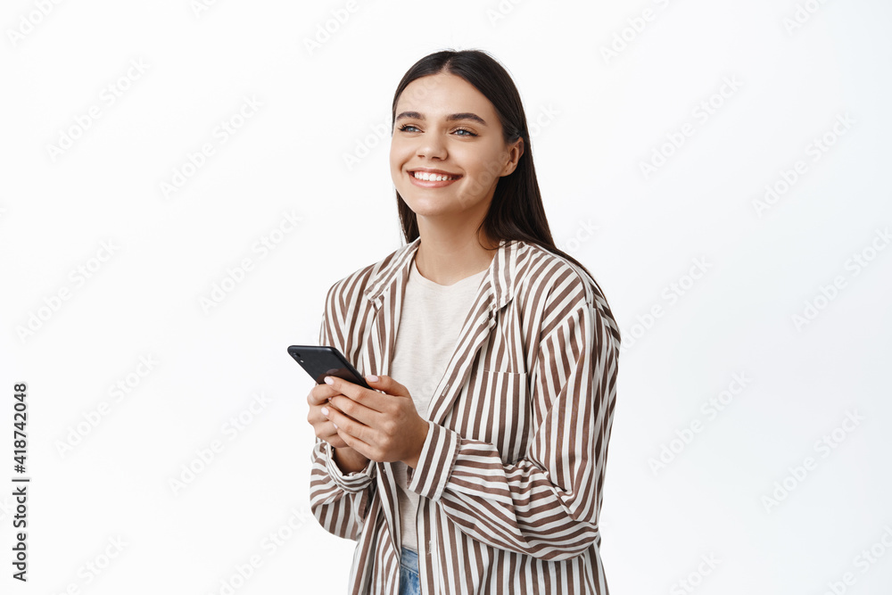 Portrait of smiling caucasian woman in trendy outfit, chatting or using app, looking happy aside, standing against white background Portrait of smiling caucasian woman in trendy outfit, chatting or using app, looking happy aside, standing against white background