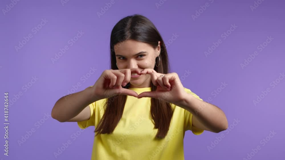 Kind pretty woman making sign of shape heart near her chest. Violet background. Women health, volunteering, donation help and love concept. Kind pretty woman making sign of shape heart near her chest. Violet background. Women health, volunteering, donation help and love concept.