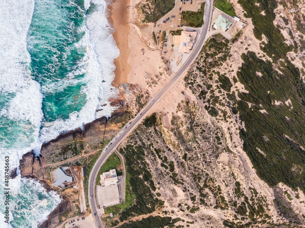 Aerial view of a narrow road with vehicles driving along the wild south coastline facing the Atlantic Ocean wit crispy waves rolling on the cliffs, Cascais, Portugal.