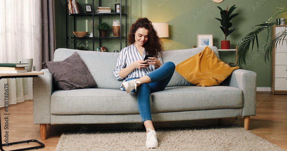 Obraz premium Caucasian pretty young cheerful smiling female sitting on sofa and texting on smartphone resting in room at home browsing online searching internet, social network user, mobile phone application