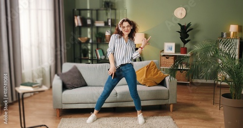 Happy joyful young Caucasian beautiful woman in positive mood dancing moving rhythmically and jumping while listening to music song in headphones on smartphone, having fun in room, leisure concept