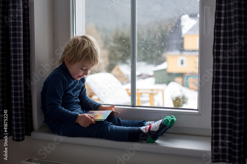 Toddler child, sitting on the window, watching the snow falling, reading a book Free Download Toddler child, sitting on the window, watching the snow falling, reading a book - GFXLoad