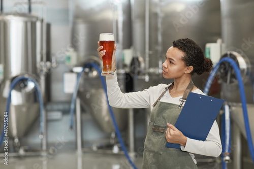 Foto Waist up portrait of young African-American woman holding beer glass while inspe