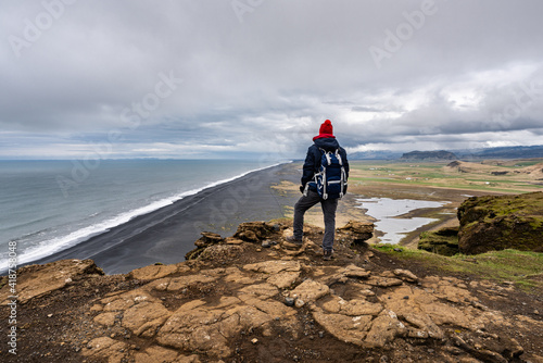 Scenery from Dyrhólaey View Point in South Iceland with a man alone standing at the edge of the cliff with a backpack in a red beanie