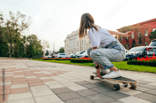 Back view on a female skater in casual attire riding her skateboard on a concrete pavement in a city park. Flowers and trees on a background.