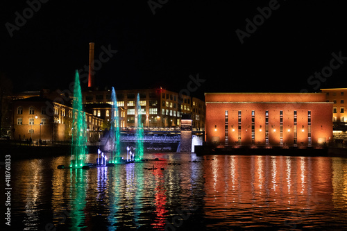 Fotografi Lighted brick buildings with red and green fountains above water in front