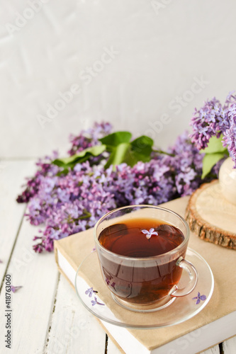 Romantic background with a cup of tea, lilac flowers and a book over a white wooden table. Leisure concept, spring breakfast