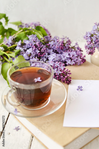 Romantic background with a cup of tea  lilac flowers and a book over a white wooden table. Leisure concept  spring breakfast. Mockup.