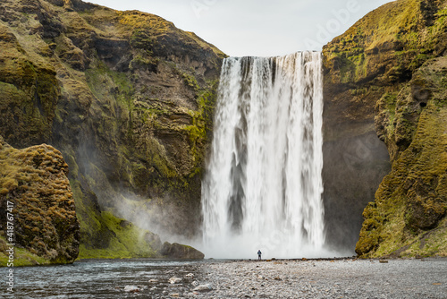 View of famous Sk  gafoss waterfall with a man standing at the waterfall alone on a sunny spring day. Popular Travel destinations. Classic Icelandic Landscape