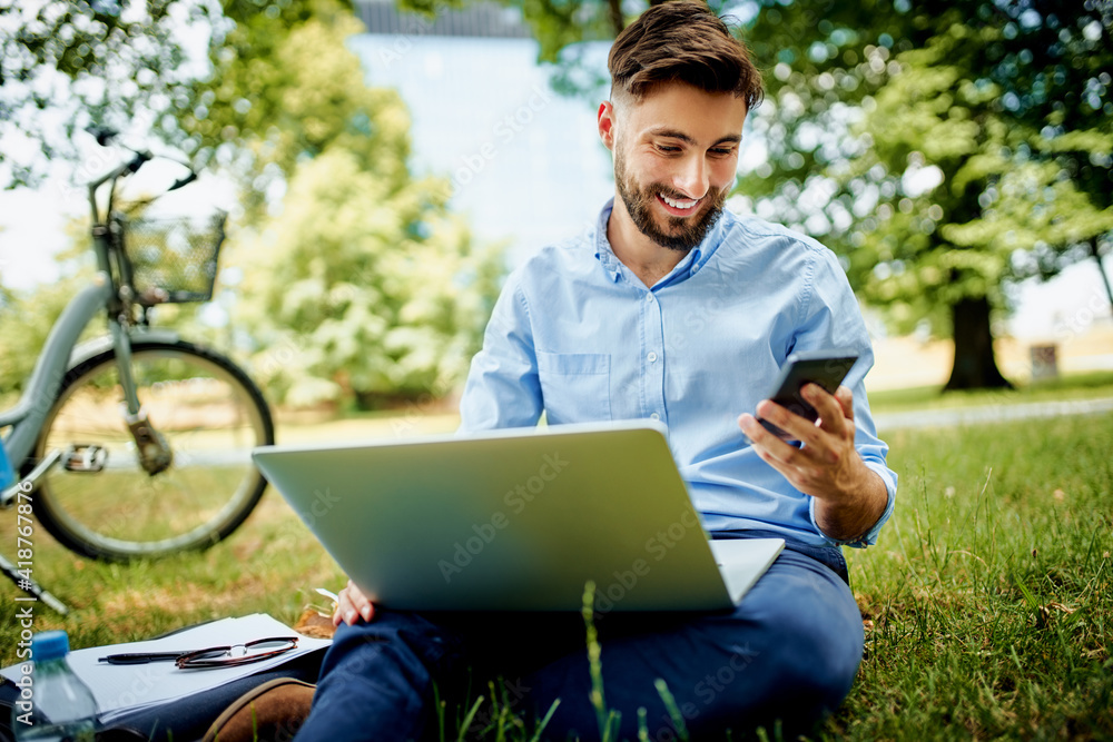 Young businessman checking phone while sitting in the park with bicycle and laptop - obrazy, fototapety, plakaty 