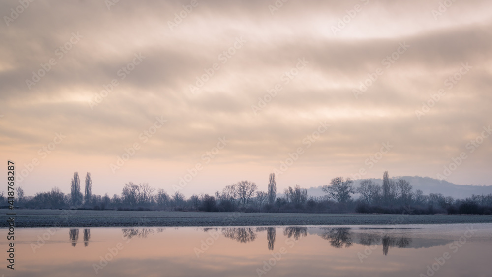 Moving clouds at dawn reflected in a small lake with trees and a hill