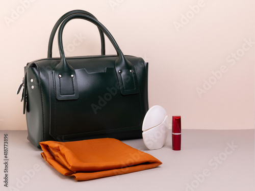 The tote bag is made of dark green smooth leather for women, with an orange neck scarf, perfume and red lipstick. Gray and cream background