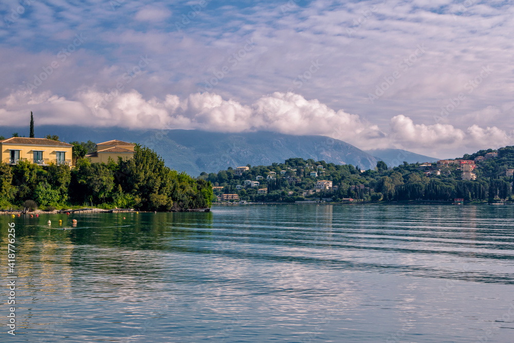 Beautiful idyllic summer landscape with calm sea, blue sky and white clouds, reflecting on water surface, green trees and mountains on the horizon. Corfu Island, Greece.