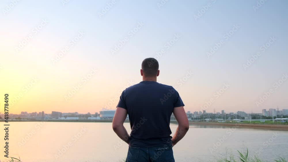 Back view of lonely caucasian man standing and enjoying view looking at lake on the center of small city and stadium. Rear view of young guy in shorts and blue t-shirt observes the surface of river. Back view of lonely caucasian man standing and enjoying view looking at lake on the center of small city and stadium. Rear view of young guy in shorts and blue t-shirt observes the surface of river.
