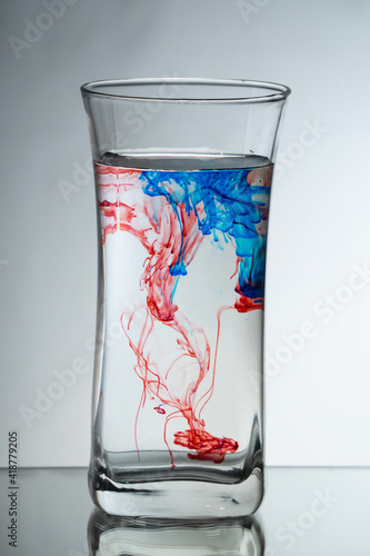 Single drops of light blue and red ink falling down into the glass full of water in the studio. Reflecting surface and gradient background