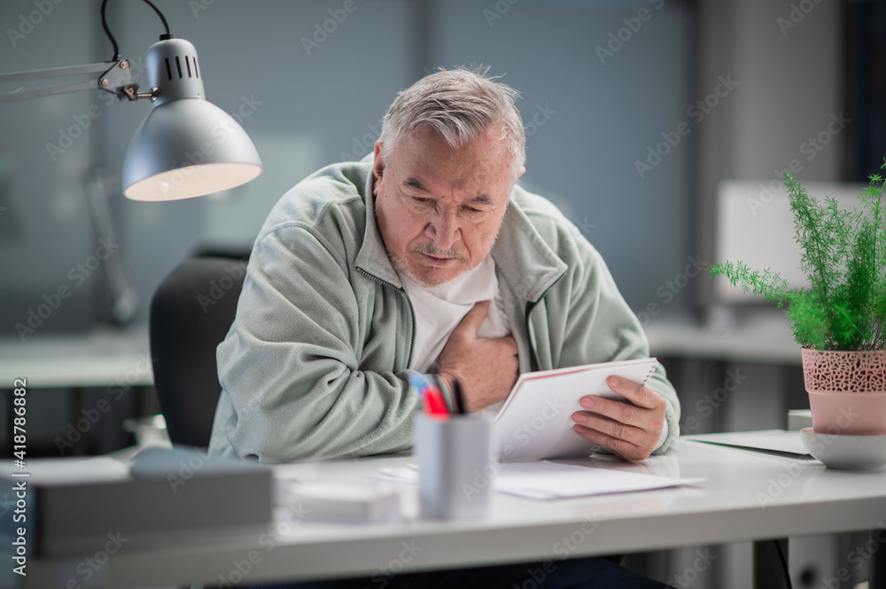 In his office, a gray-haired man felt a pain in his heart while working, he sits with his hand on his chest. In his office, a gray-haired man felt a pain in his heart while working, he sits with his hand on his chest.