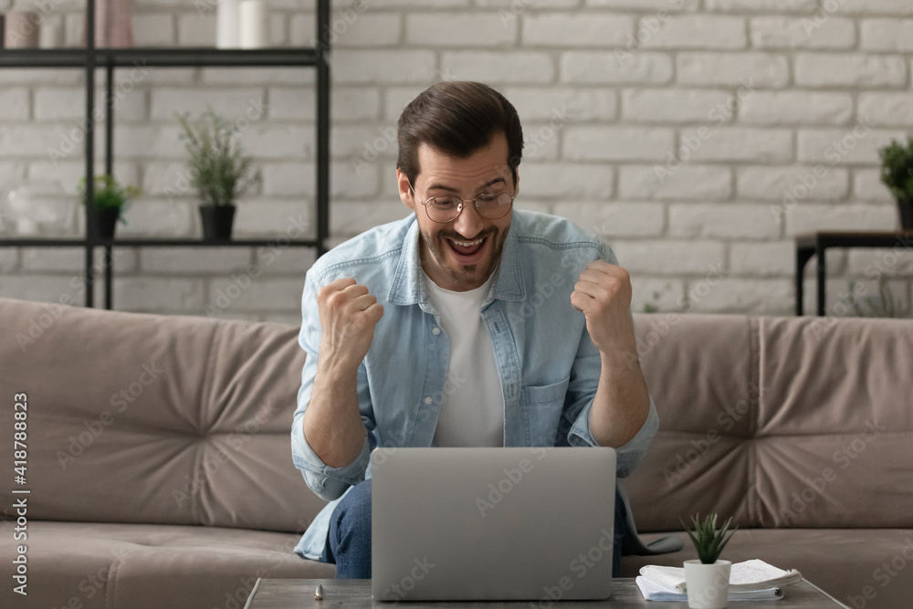 Great thing. Euphoric young man sit on couch by laptop screen shout super raise fists in winner gesture get good news job offer email. Amazed excited funny guy celebrate victory in web game of chance Great thing. Euphoric young man sit on couch by laptop screen shout super raise fists in winner gesture get good news job offer email. Amazed excited funny guy celebrate victory in web game of chance