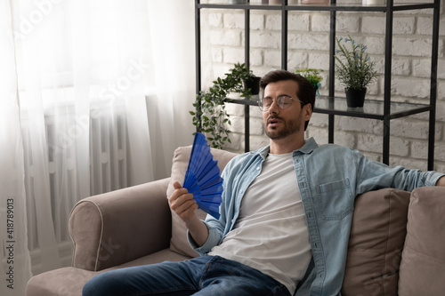 Frustrated guy recline on sofa suffer from too hot temperature at home breath hard with dehydrated air cool down with hand fan. Tired young male complain on extra heat at living room feel bad sweaty