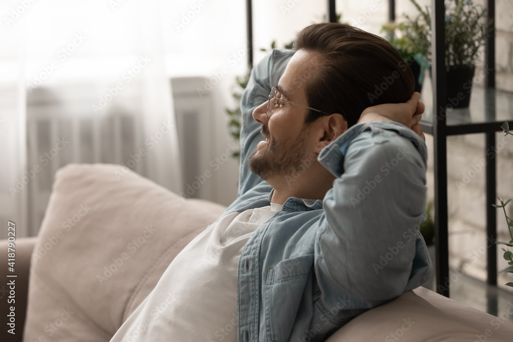 Side shot of happy tranquil millennial man in glasses sit on cozy sofa hold hands behind head look aside relax dream. Calm young male rest at home in meditative mood take break from work. Copy space Side shot of happy tranquil millennial man in glasses sit on cozy sofa hold hands behind head look aside relax dream. Calm young male rest at home in meditative mood take break from work. Copy space