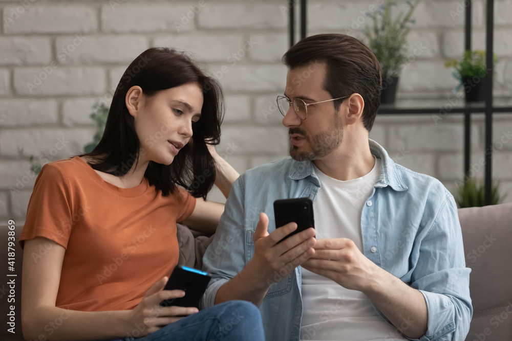 Young family couple sit on couch hold smartphones discuss work of new web app share information between gadgets. Diverse friends man woman users of modern phones talk send receive get photo video file Young family couple sit on couch hold smartphones discuss work of new web app share information between gadgets. Diverse friends man woman users of modern phones talk send receive get photo video file