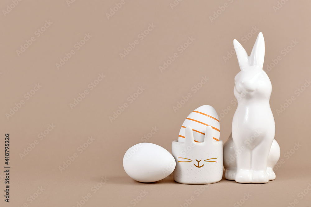 Golden striped Easter egg in bunny egg cup next to rabbit figure and white egg on side of beige background with empty copy space Golden striped Easter egg in bunny egg cup next to rabbit figure and white egg on side of beige background with empty copy space