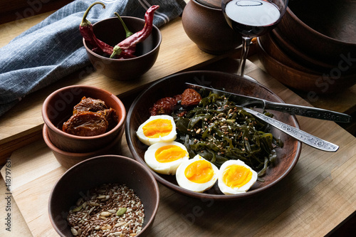 Boiled eggs, seaweed, sun-dried tomatoes sprinkled with pumpkin seeds, flax seeds and seeds in a clay plate and a glass of wine on a light wooden surface