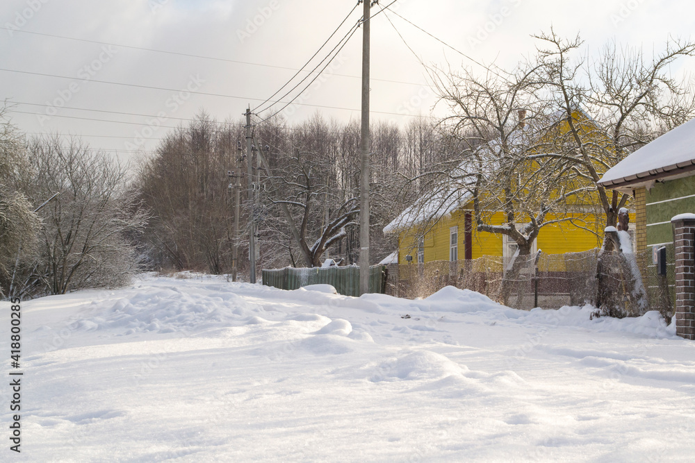 A cloudy winter day with snowfall. A street in a dacha village. Snow covered rural road and snowdrifts along the road near the fences.