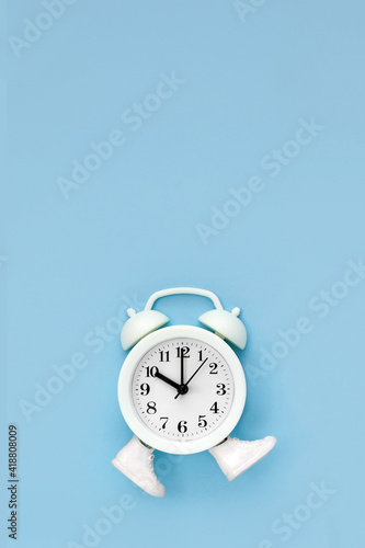 White alarm clock in toy sneakers walks on blue background. Time movement concept. Vertical poster, copy space