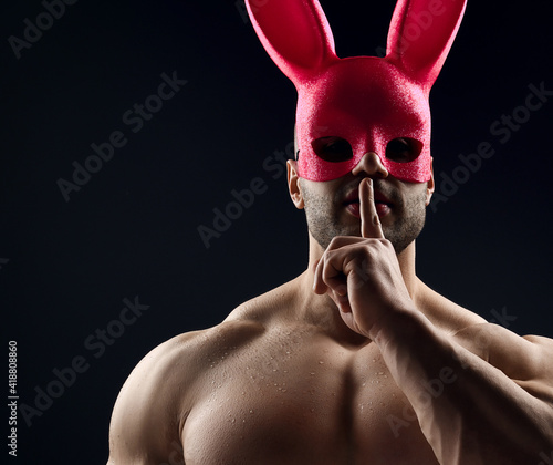 Slika na platnu Brutal sweaty strong young man with naked pumped up upper body in pink rabbit mask standing showing silence sign with fingers over grey background
