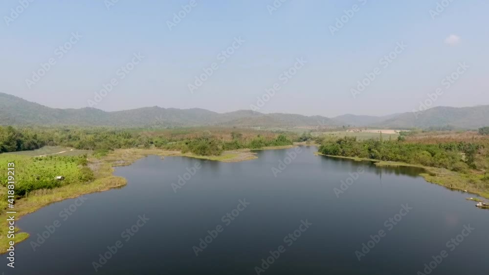 The aerial view of the blue calm lake surrounded by the fields and the forest under the strong sunlight and the blue clear sky with the mountains in the background The aerial view of the blue calm lake surrounded by the fields and the forest under the strong sunlight and the blue clear sky with the mountains in the background