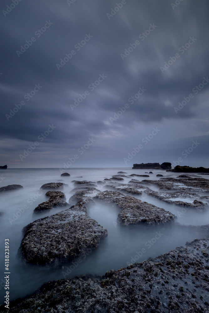 Obraz premium Dreamy seascape for background. Beach with rocks and stones. Low tide. Blurred foggy water. Cloudy sky. Slow shutter speed. Soft focus. Copy space. Mengening beach, Bali
