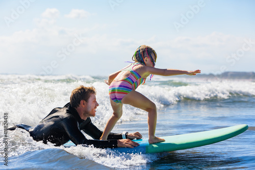 Little surf girl - young surfer learn to ride on surfboard with instructor at surfing school. Active family lifestyle, kids water sport lessons, swimming activity in summer camp. Vacation with child.