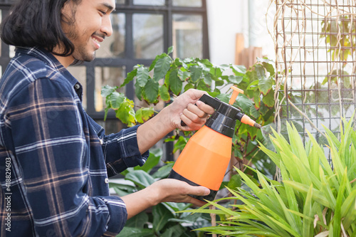 Smiling, happy young asian man, gardener is holding a water sprayer to water the plants with spray in free time activities, sunshine in the morning at home,apartment.Lifestyle, hobby people concept.