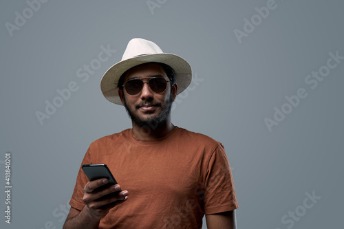 Self confident and bearded indian guy in orange shirt poses in gray background holding his smartphone. © Fxquadro