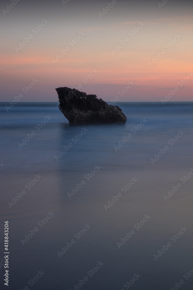 Sunset seascape. Rock in the ocean. Slow shutter speed. Soft focus. Silky water. Colorful sky. Amazing water reflection. Nature and environment background. Copy space. Beach in Bali.