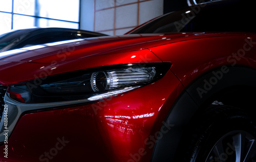 Closeup headlight of shiny red luxury SUV car in showroom. Elegant electric car with sport design. Car parked in showroom. Car dealership. Electric vehicle development concept. Future transportation. © Artinun