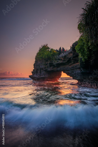 Natural arch. Batu Bolong temple on the rock during sunset. Seascape background. Motion milky waves on black sand beach. Copy space. Vertical layout. Tanah Lot, Bali
