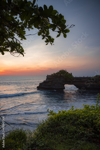 Natural arch. Batu Bolong temple on the rock during sunset. Seascape background. Motion waves. Foreground with tree branches. Copy space. Vertical layout. Tanah Lot, Bali