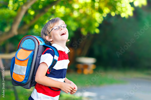 Happy little kid boy with glasses and backpack or satchel on his first day to school or nursery. Child outdoors on warm sunny day, Back to school concept.
