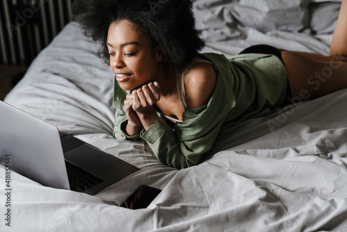 Happy african american girl using laptop while lying in bed after sleep