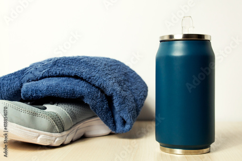 Selective focus of a blue water bottle placed on a wooden platform with sneakers covered by a towel on the background. Concept of working out indoors, being active, losing weight.