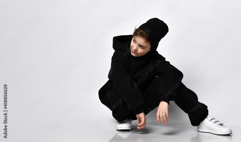 Stylish kid bro squatting on against a white wall. He is wearing a trendy black hoodie and sweatpants. black beanie and white sneakers Stylish kid bro squatting on against a white wall. He is wearing a trendy black hoodie and sweatpants. black beanie and white sneakers