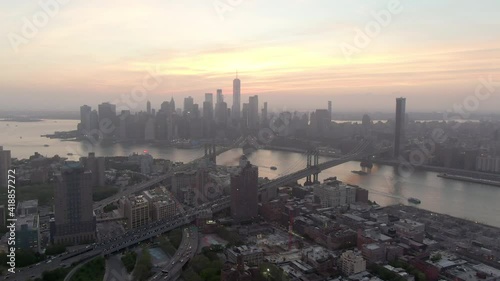 Aerial shot of vehicles and buildings in city by East River, drone flying forward towards Manhattan against sky during sunset - New York City, New York