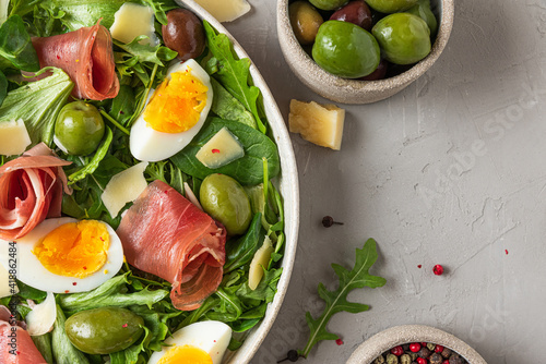 Prosciutto di Parma salad with olives, parmesan cheese, eggs and arugula on gray background. Italian appetizer. Top view