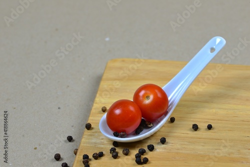 Two small red tomatoes on an Asian porcelain spoon. Light brown background with a wooden board and balls of black pepper. Decoration of fresh vegetables, spices, white spoon and wood. Ready to cook.