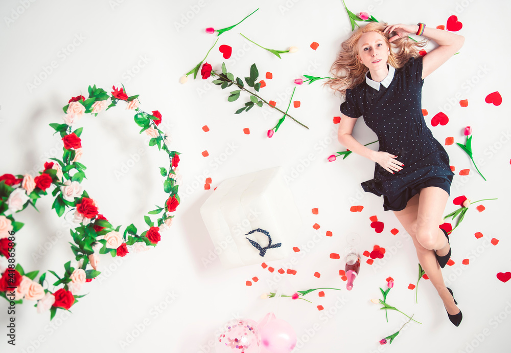Fototapeta premium Beautiful woman in a black dress and high heels lies on a white floor with a flowers on International Women's Day.