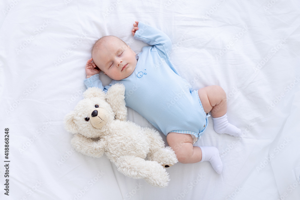 baby boy sleeps on the bed lying on his back with a soft toy bear in blue pajamas holding his hands up, healthy newborn sleep baby boy sleeps on the bed lying on his back with a soft toy bear in blue pajamas holding his hands up, healthy newborn sleep
