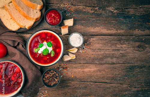 Culinary background with space to copy. Beetroot soup with sour cream and basil among garlic, spices and bread on a wooden background.